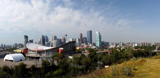 Panoramic view of Calgary skyline with tall buildings and the Scotiabank Saddledome under a partly cloudy sky.