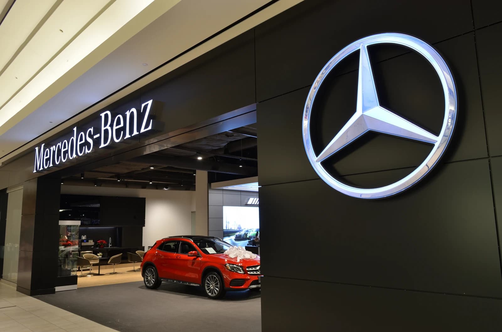 Mercedes-Benz dealership exterior with large illuminated logo and red car on display inside.