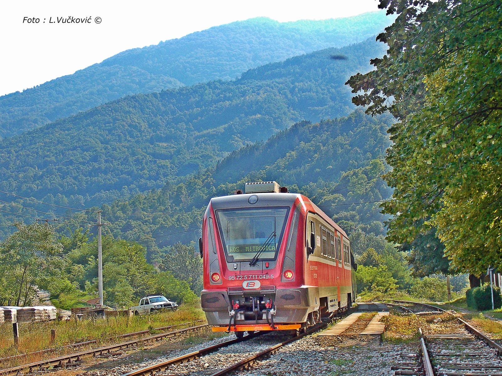 A red passenger train on a single track with mountains and trees in the background.
