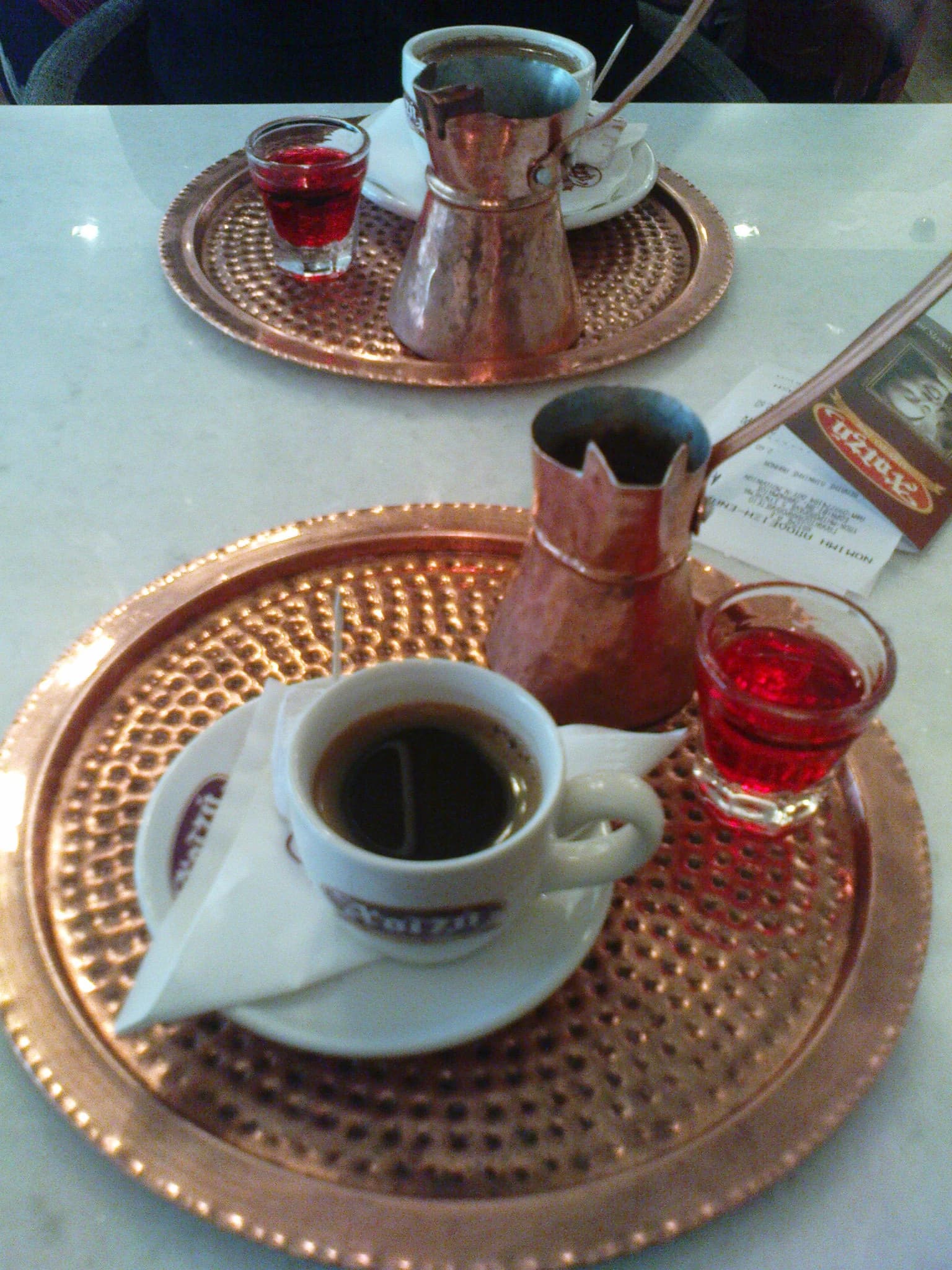Two copper trays, each with a cup of coffee, a small copper pot, and a shot of red liquid.