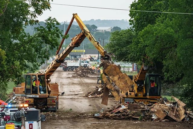 Two Gradall excavators with operators moving large debris from a muddy street.