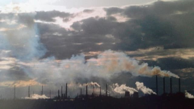 Silhouette of PCK Schwedt refinery with smoke plumes rising against a cloudy sky.