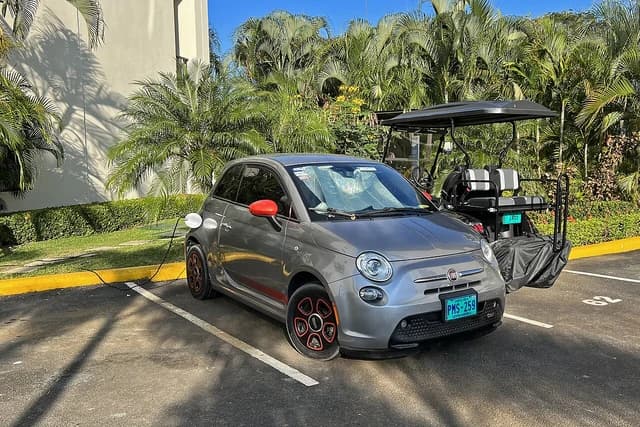 A grey Fiat 500e electric car is parked and charging in a sunny parking lot.