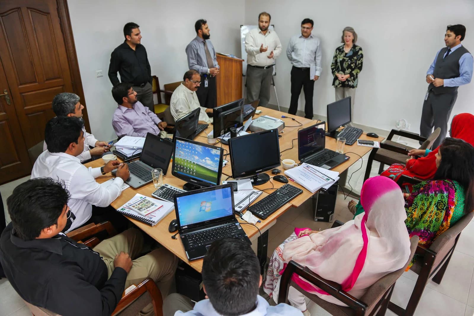 Group of men and women attending a computer skills training class, some using laptops.