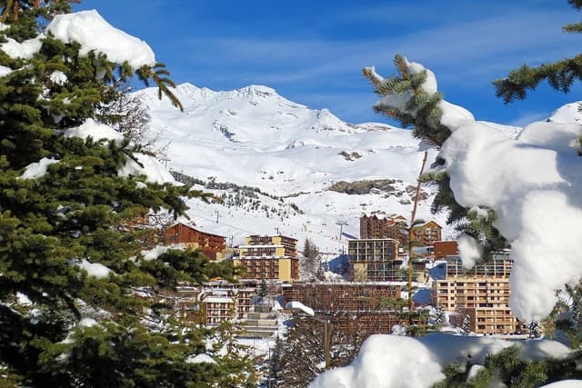 Ski resort buildings among snow-covered trees and mountains under a blue sky.