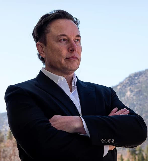Elon Musk, CEO of Tesla, stands outdoors with arms crossed.