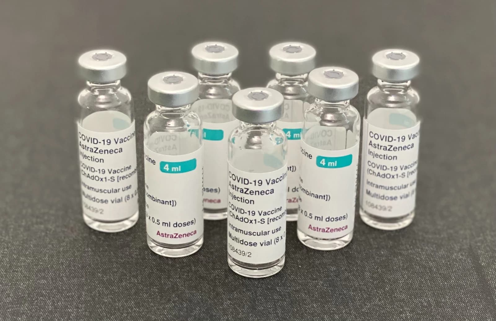 Several clear glass vials containing AstraZeneca COVID-19 vaccine on a dark surface.