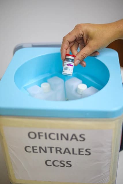 A hand holds a vial of AstraZeneca vaccine above a cooler labeled 'Oficinas Centrales CCSS'.