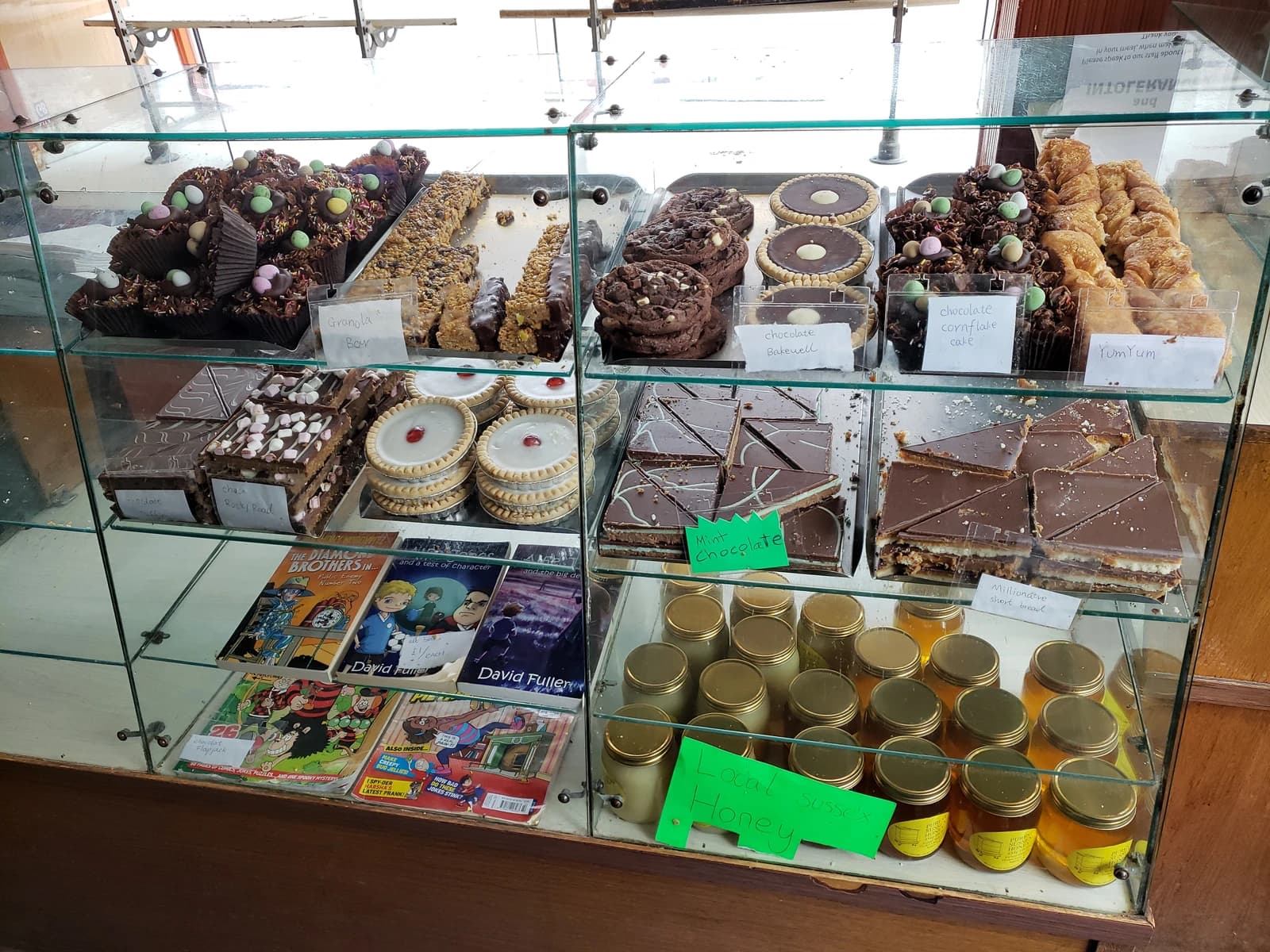 A glass display case shows rows of various baked goods, honey jars, and books for sale.