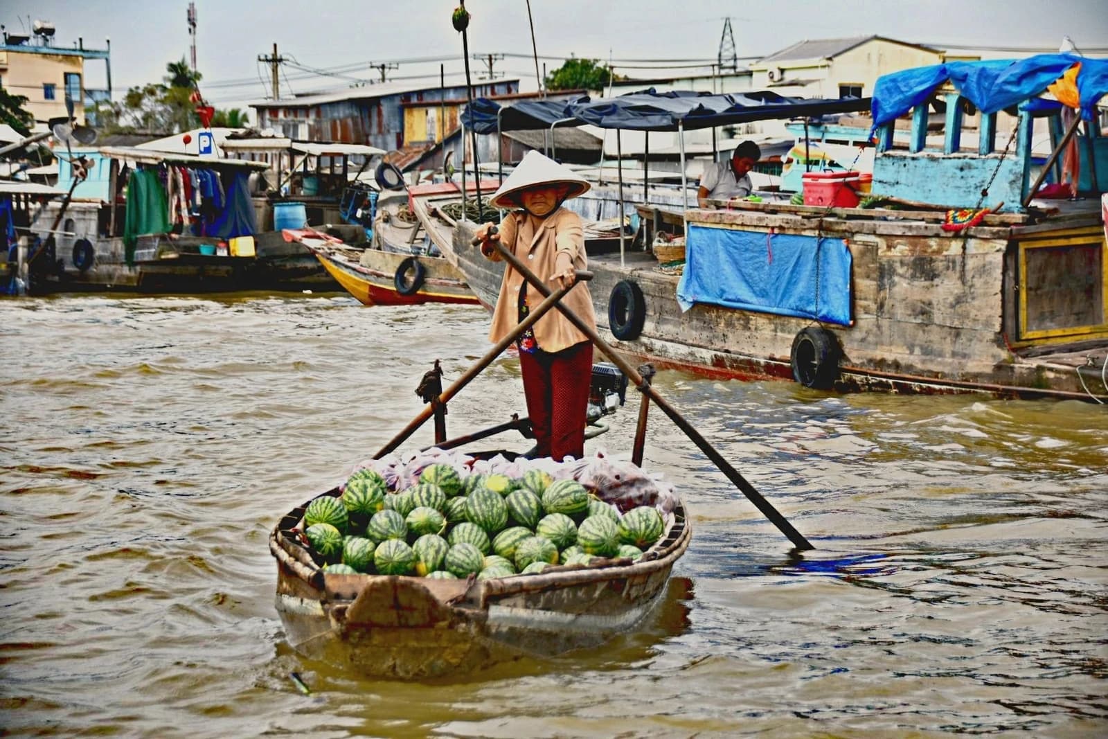 Woman in a conical hat paddling a small boat loaded with watermelons on a river.