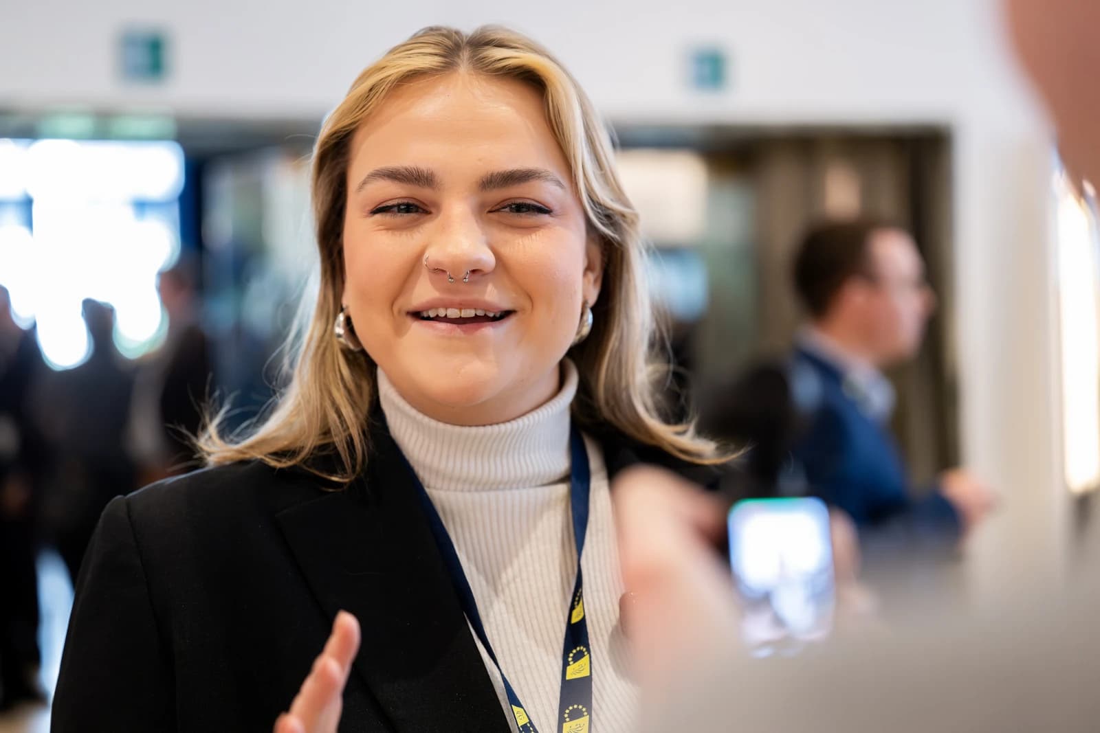 Young woman with blonde hair smiling, wearing a white turtleneck and black blazer.