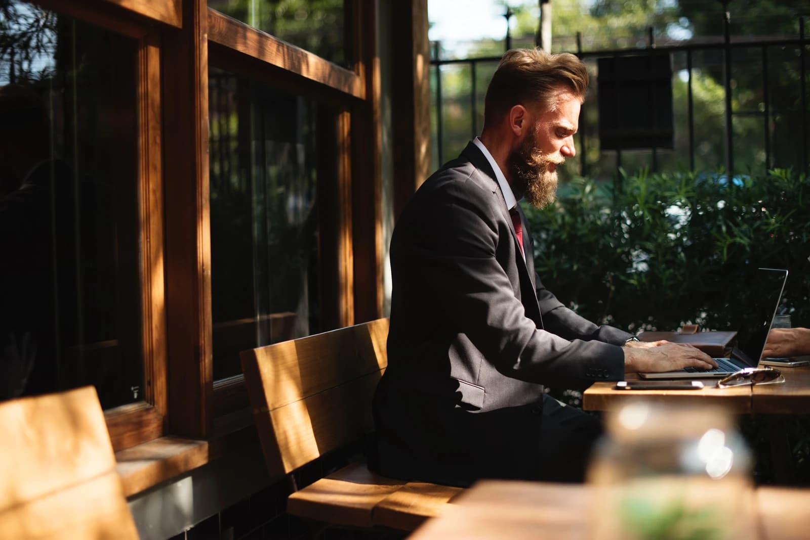 Man with beard typing on a laptop computer at an outdoor cafe table.