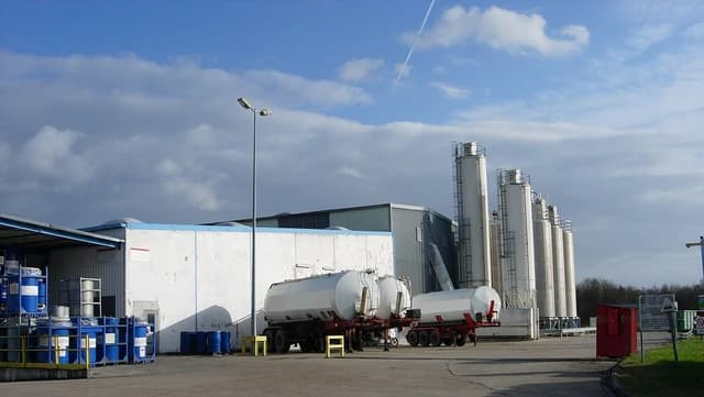An industrial facility with large white silos and tanker trucks under a partly cloudy sky.