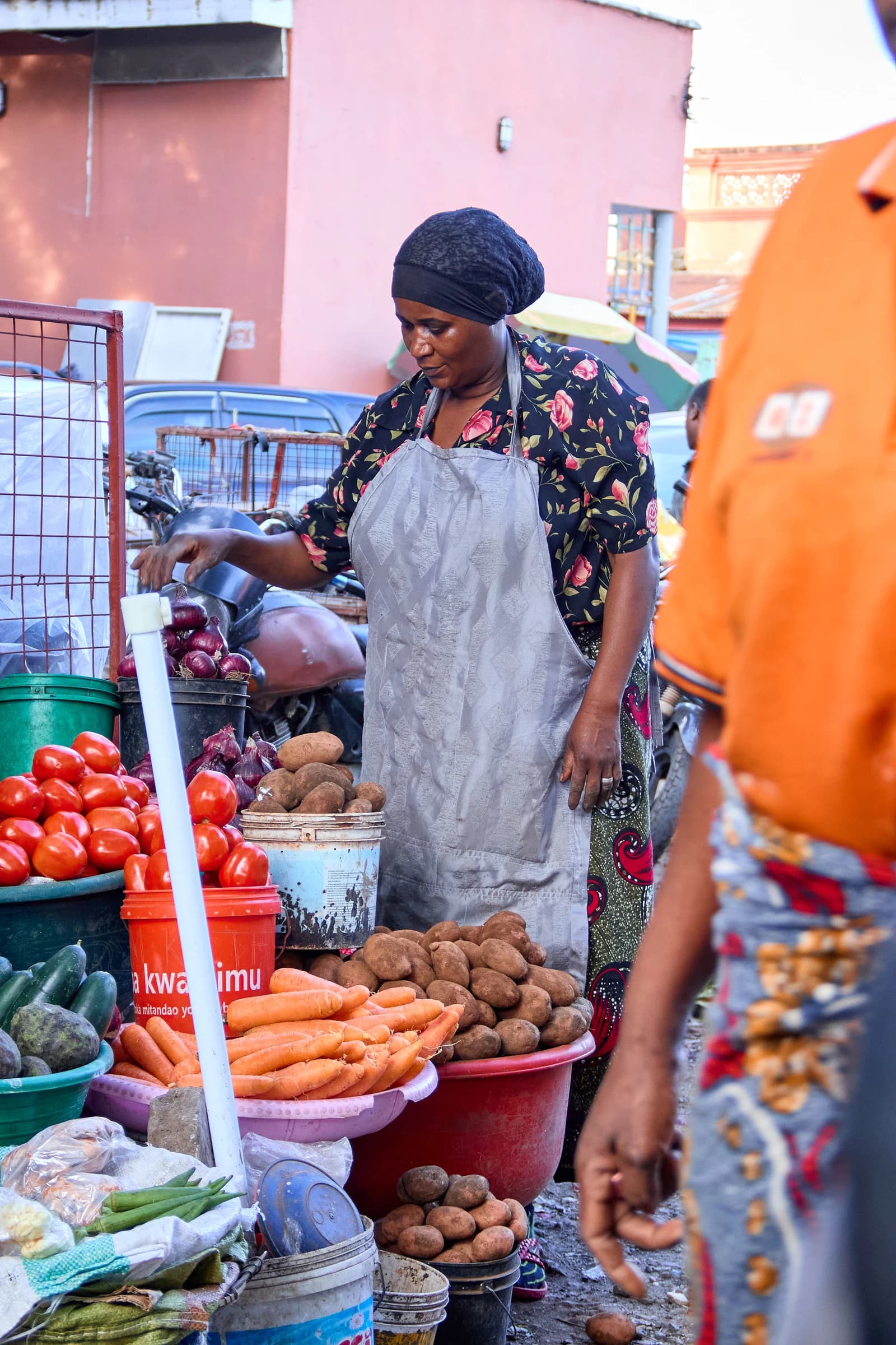 A market vendor in an apron arranging fresh tomatoes, potatoes, onions, and carrots on a stall.