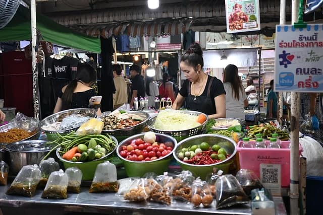Vendor prepares dishes behind counter at night market food stall displaying fresh vegetables.