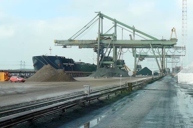 Bulk carrier ship 'Pacific Resource' at a port, with large gantry cranes and piles of materials.