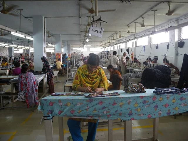 Women operate sewing machines and sort textiles in a brightly lit garment factory.