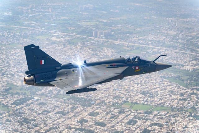 An Indian Air Force Tejas MK1 fighter jet flying over a city with a pilot in the cockpit.