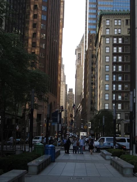 Pedestrians walk on a sidewalk lined with buildings on Wall Street in Manhattan.