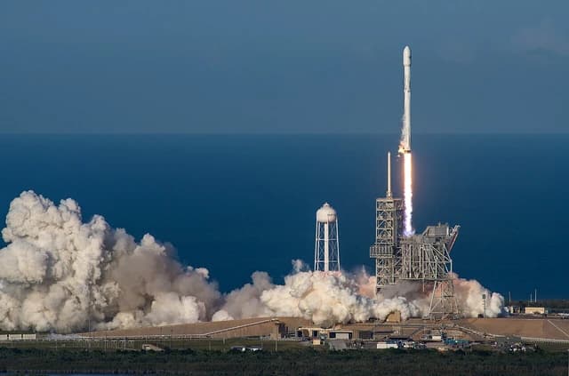 SpaceX Falcon 9 rocket lifting off from launchpad with ocean in background.