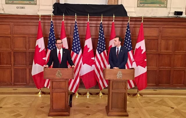 Steven Mnuchin and Bill Morneau stand at podiums with Canadian and American flags.