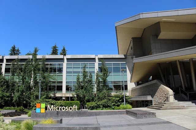 Microsoft logo sign stands in front of a modern corporate building on a clear day.