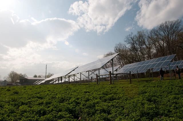 Large array of ground-mounted solar panels in a green field under a partly cloudy sky.