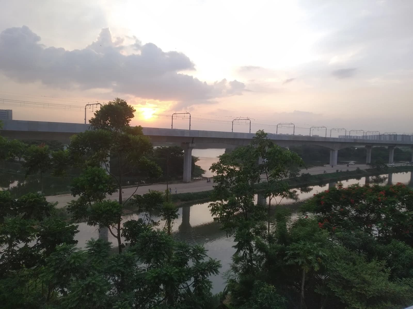 Elevated metro rail line extends over a river with trees on the banks at sunset.