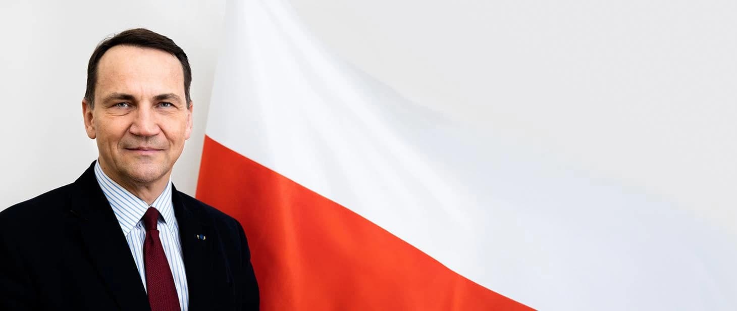 Radosław Sikorski, Polish Minister of Foreign Affairs, smiling in front of a Polish flag.