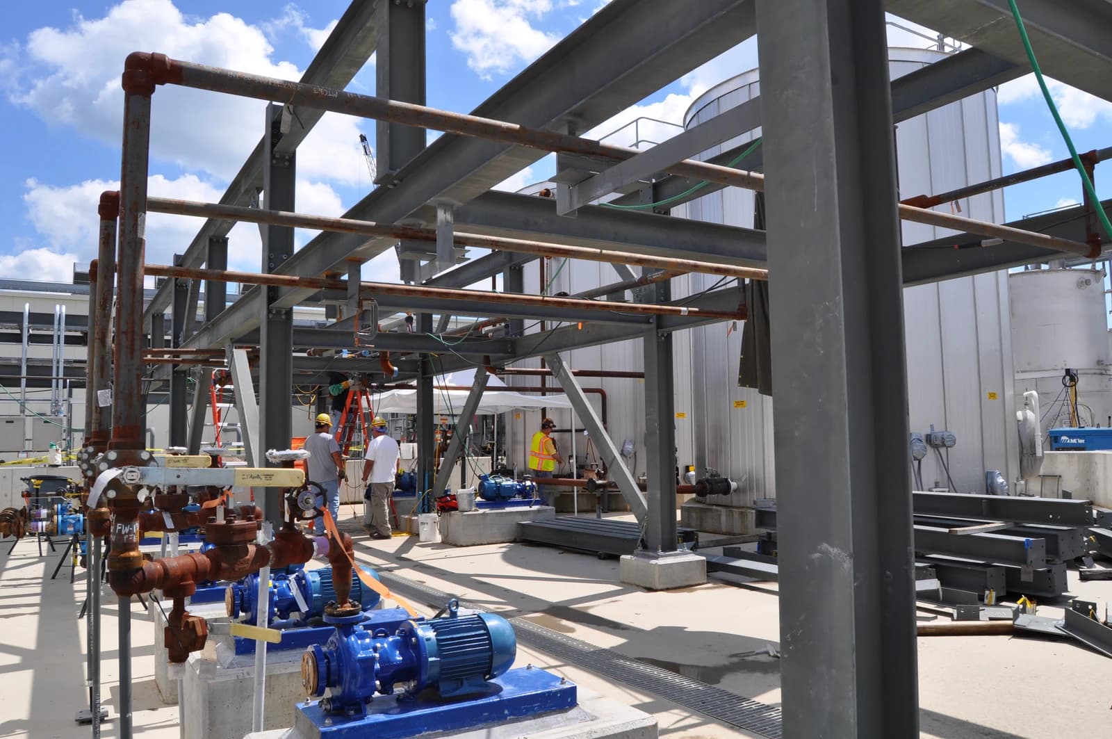 Industrial facility with pipes, pumps, and large tanks being constructed by workers.