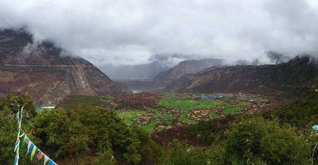 Overcast sky above a green valley, mountains, and small blue-roofed buildings in Tibet.