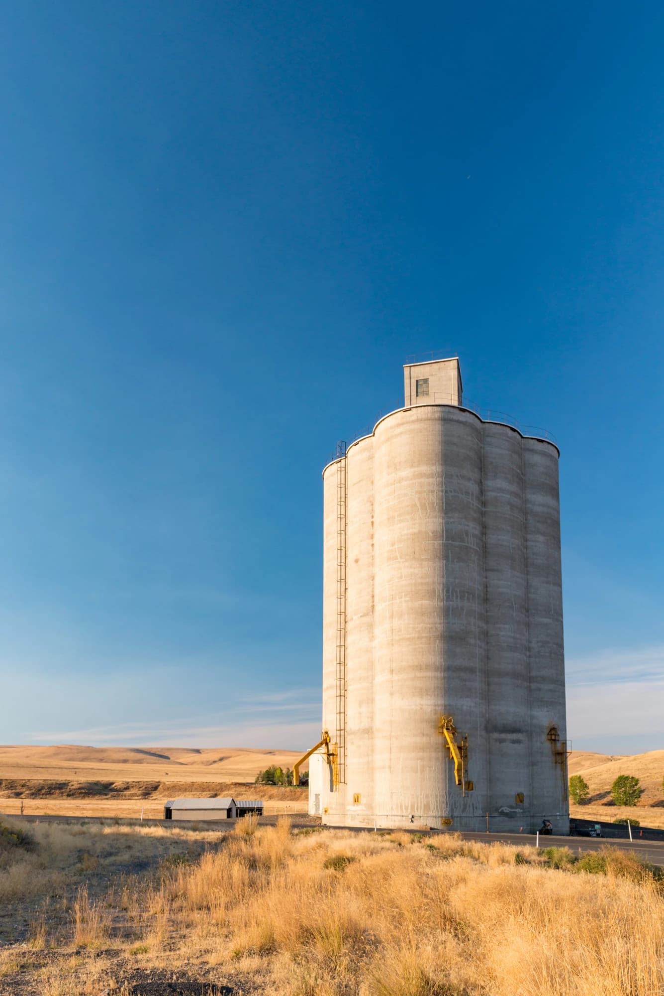 Grain silo stands tall in a dry field with a road and hills in the background.