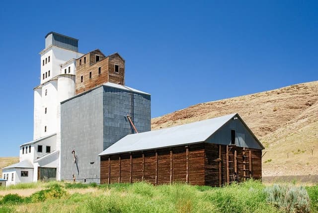 Grain elevator complex with tall white and tin-sided buildings next to a wooden barn.