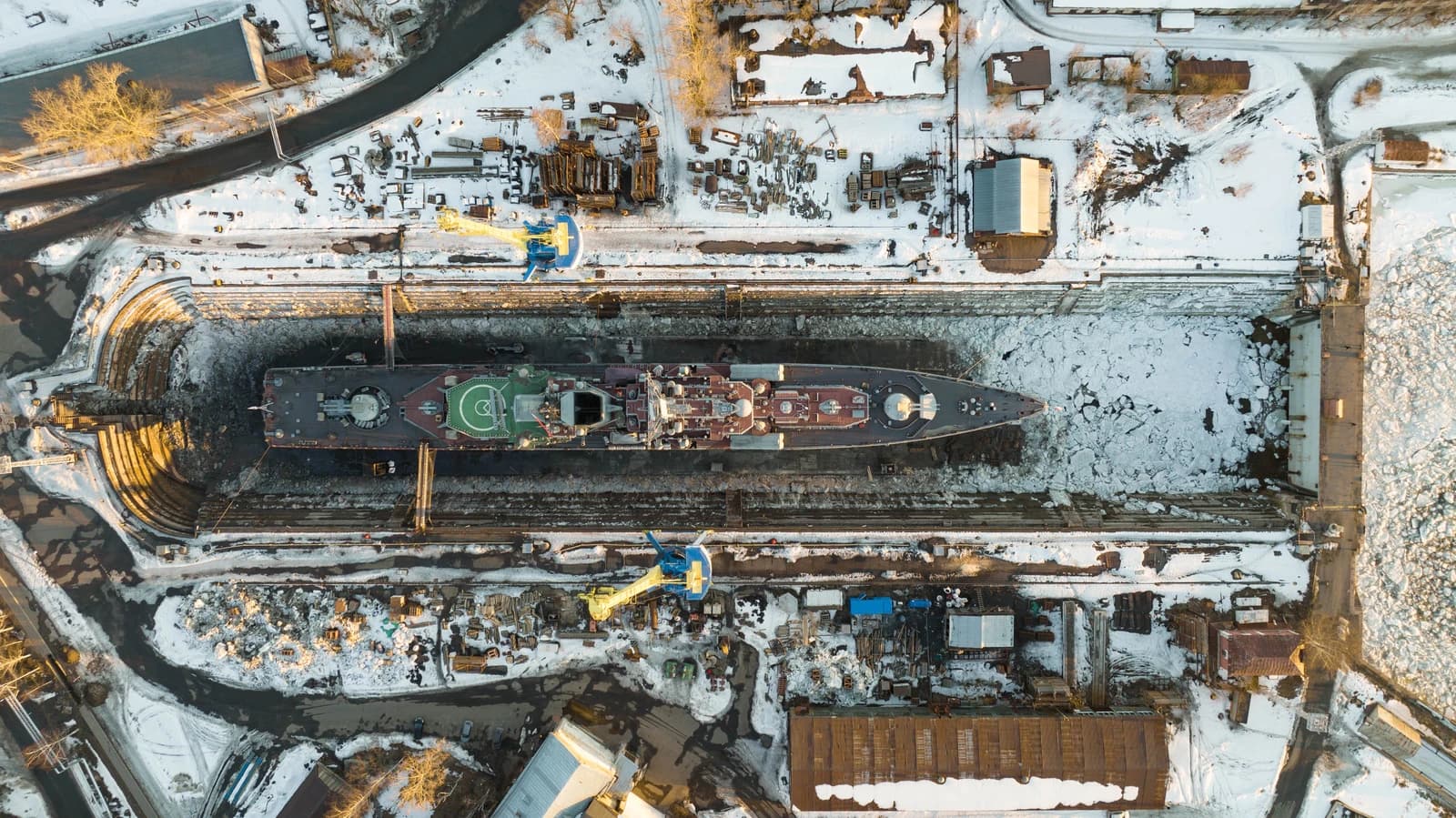 Aerial view of a warship inside a dry dock at a large industrial shipyard covered in snow and ice.