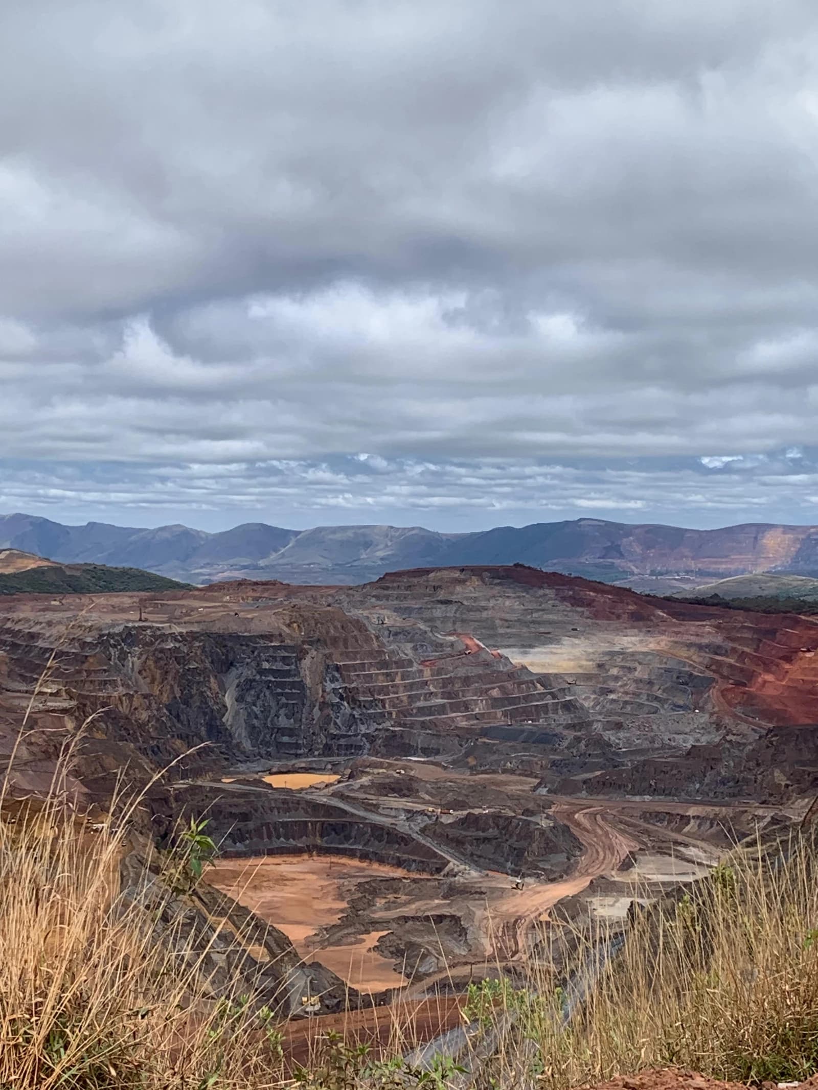 Open-pit mine with stepped terraces, ponds, and distant mountains under a cloudy sky.