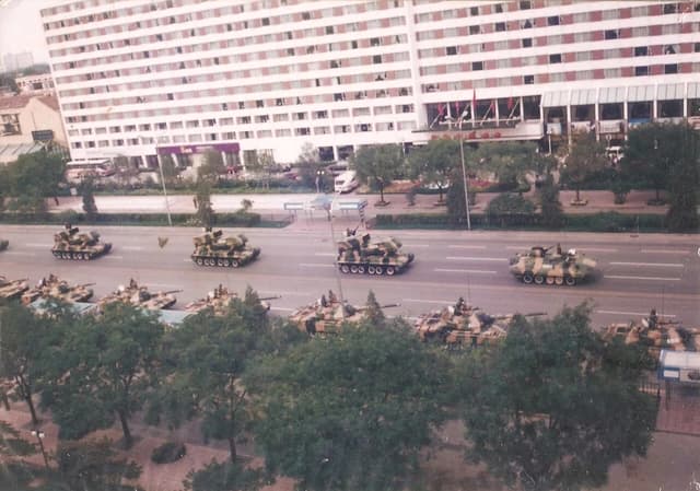 Military tanks and missile launch vehicles parade on a wide city street.