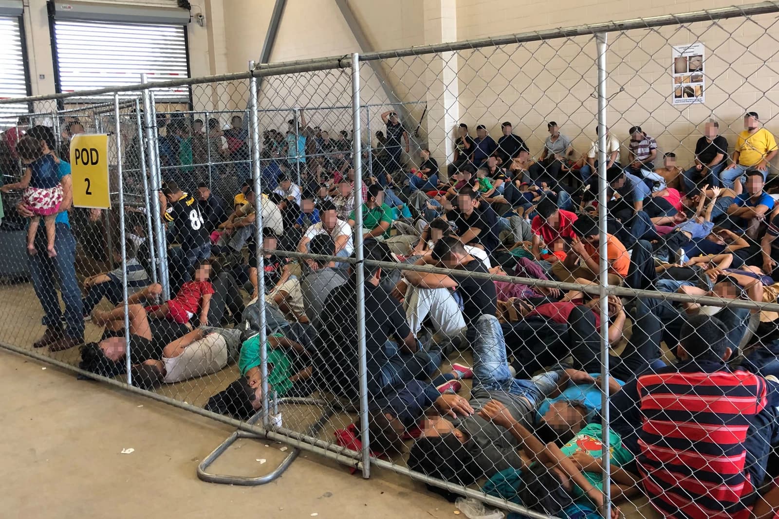 Migrants, including adults and children, resting on the floor inside an overcrowded chain-link enclosure.