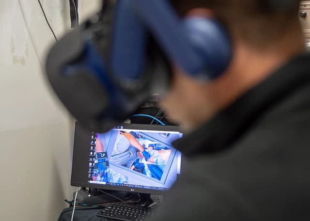 A person wears a virtual reality headset while looking at a computer screen showing surgery.