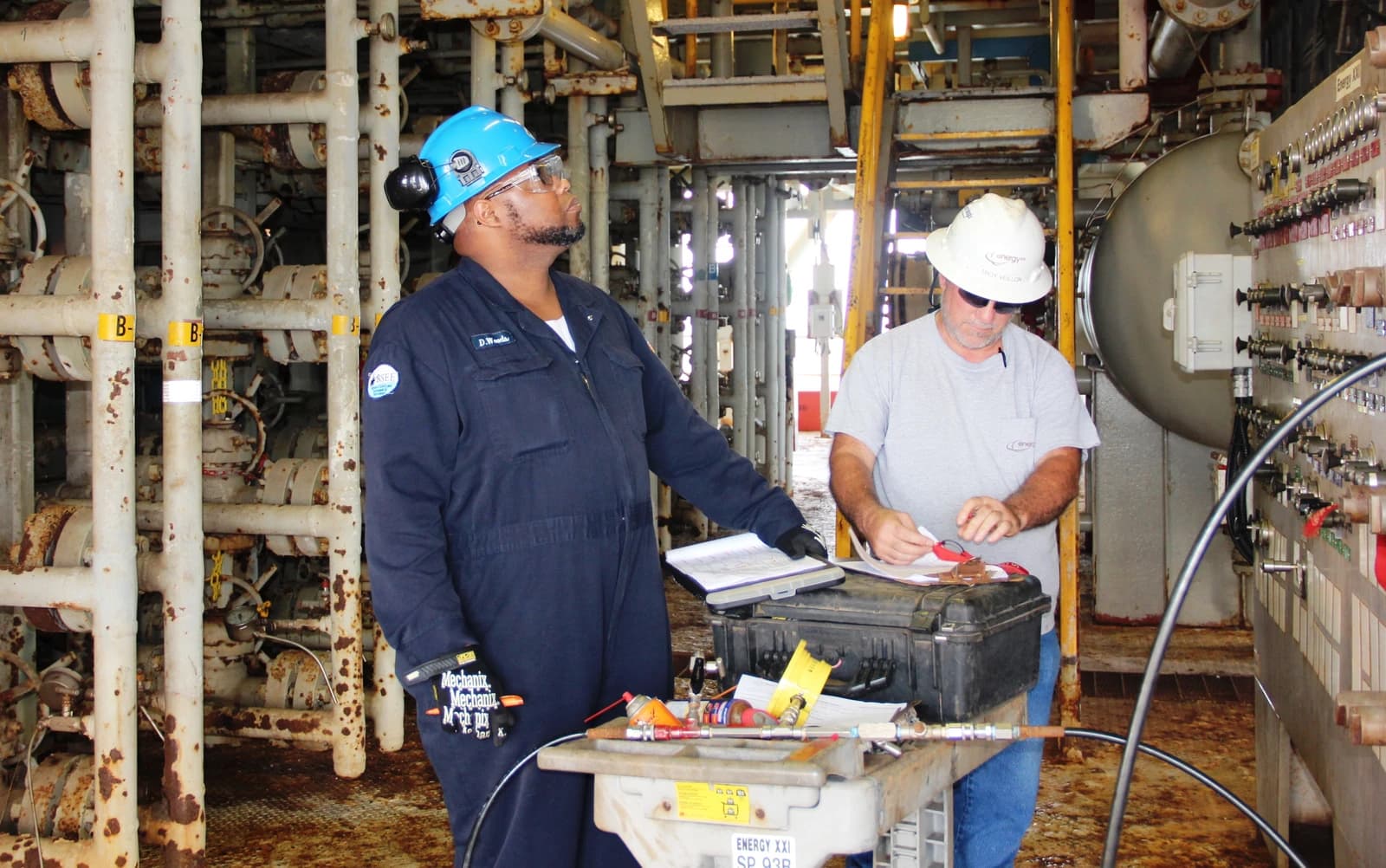 Two men wearing hard hats and safety gear working on industrial equipment.