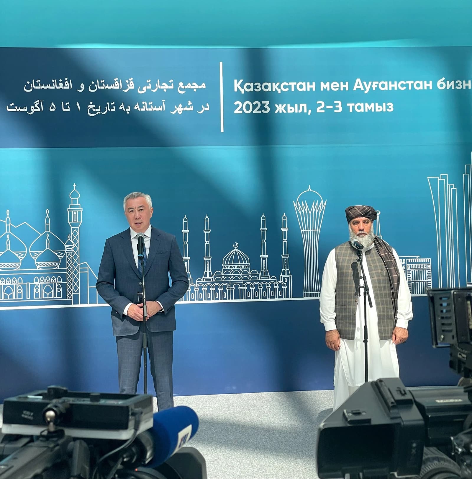 Serik Zhumangarin and Nooruddin Azizi stand at microphones in front of a blue backdrop.