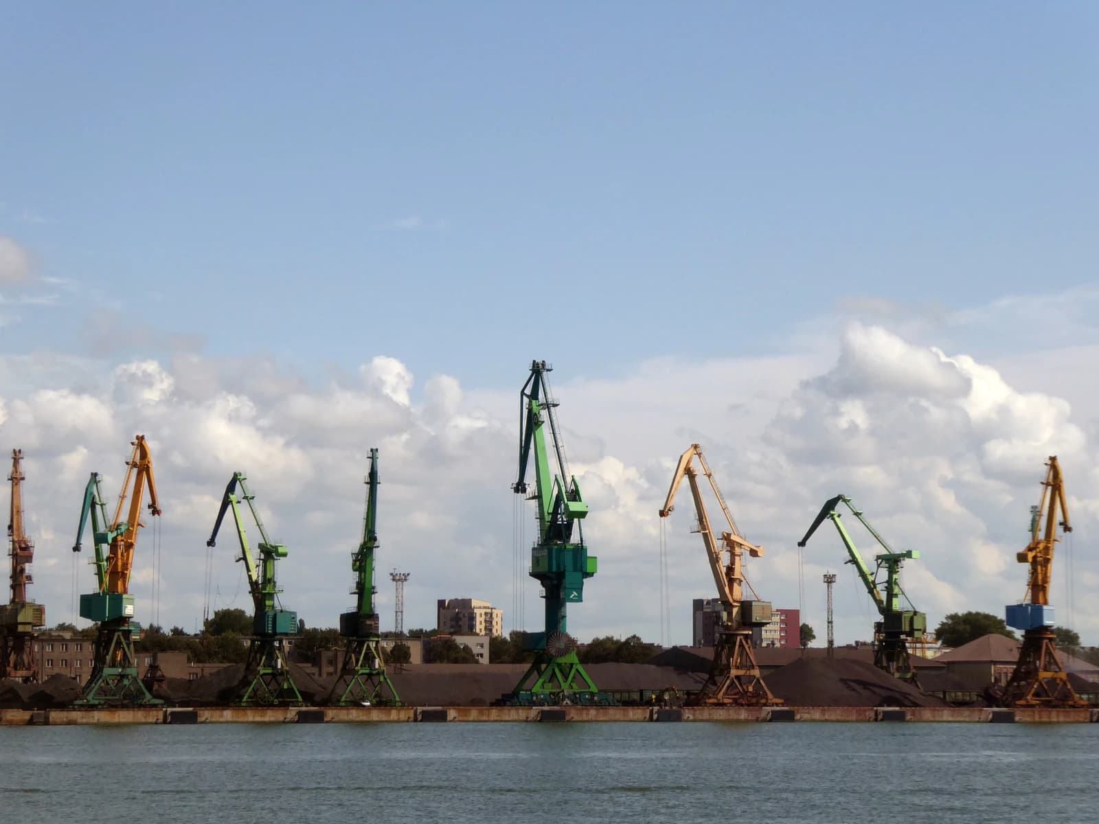 Numerous port cranes stand along a waterfront, with large piles of grain visible behind them.