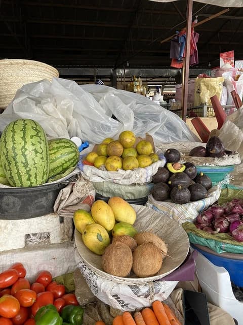 Watermelons, oranges, avocados, mangoes, coconuts, tomatoes, and carrots are displayed at a market stall.