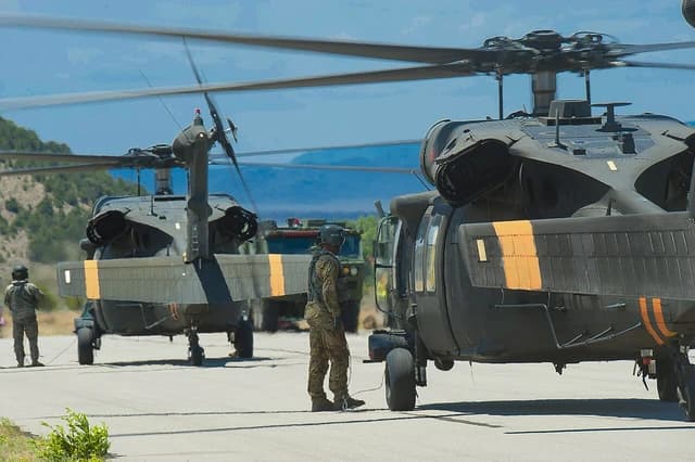 Military personnel stand on an airfield next to two Black Hawk helicopters, with hills in the background.