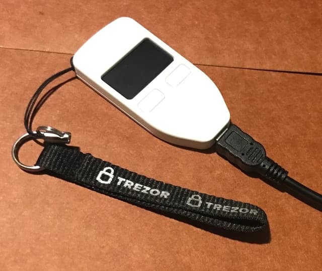 A white Trezor hardware wallet connected by a black cable, resting on a brown surface.