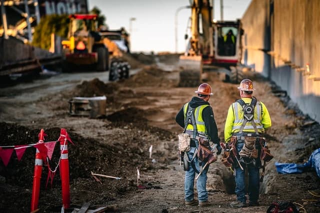 Two construction workers wearing safety helmets and vests stand on a job site.