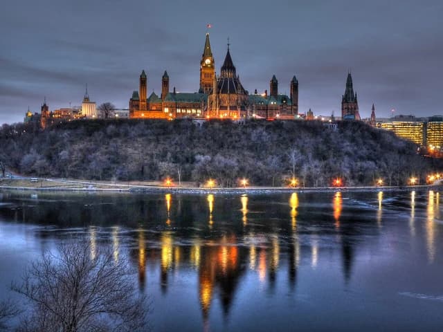 Parliament Hill buildings lit up at dawn, reflecting on the Ottawa River.