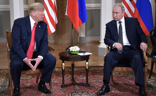 Donald Trump and Vladimir Putin sit facing each other with national flags behind them.