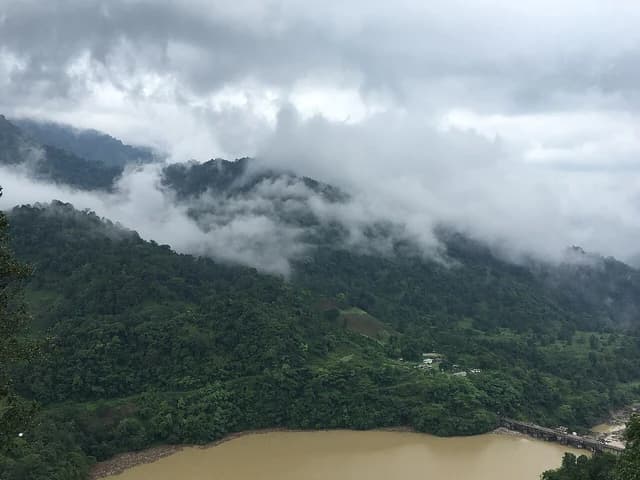 Lush green mountains shrouded in clouds beside a muddy river with a distant bridge.