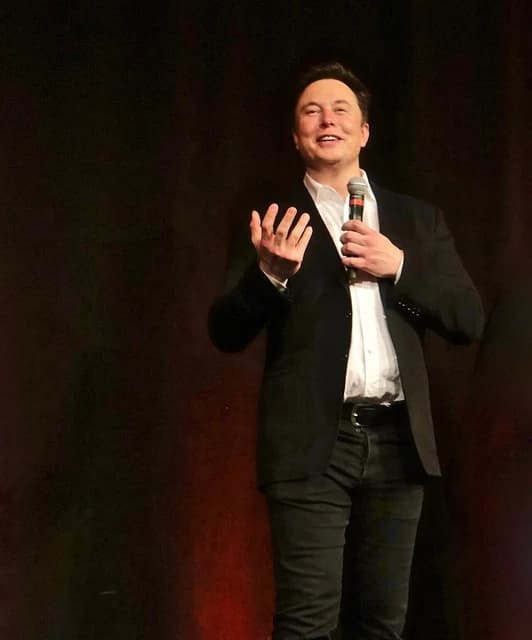 Elon Musk, CEO of Tesla, holding a microphone and gesturing while speaking.