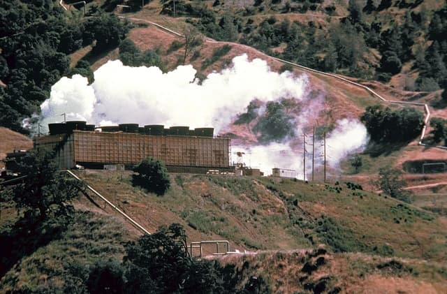 Geothermal power plant facility with steam rising from cooling towers located in a hilly, rural area.